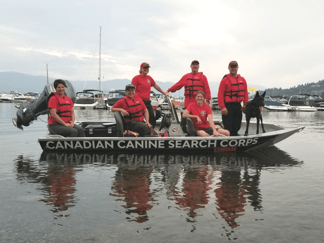 Team of 6 people in red lifejackets on a black motorboat with "Canadian Canine Search Corps" in white print. Black dog on the front of the boat.